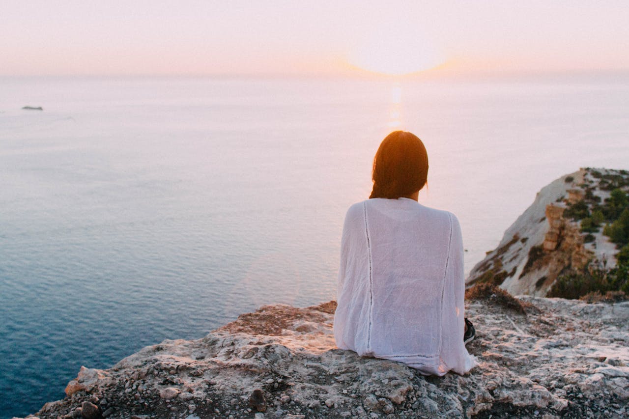 portfolio-04 A serene view of a woman sitting on a rocky cliff, gazing at the ocean during sunset. Perfect for relaxation and travel themes.